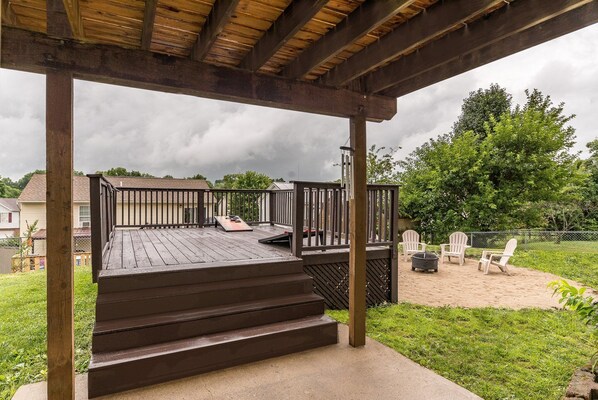 Una terraza de madera con escalones que conducen a una zona cubierta, rodeada por un patio cercado con zona de arena y césped verde.