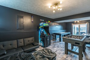 Relax in a stylish room with a sofa, foosball table, and retro arcade machine, featuring a unique ceiling design and a window with a view.