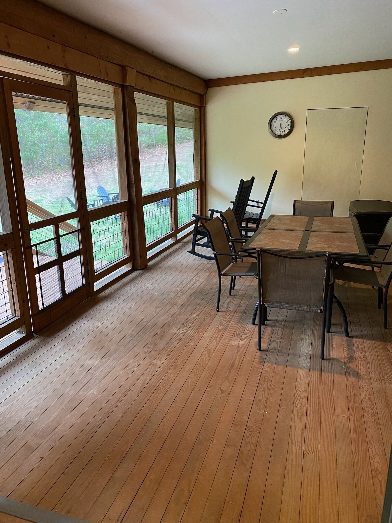 A dining area with a wooden table and chairs, large windows, and a clock on the wall.