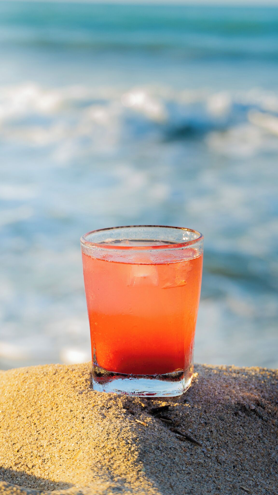 A glass of red drink on a sandy surface with a blurred ocean and sky in the background.