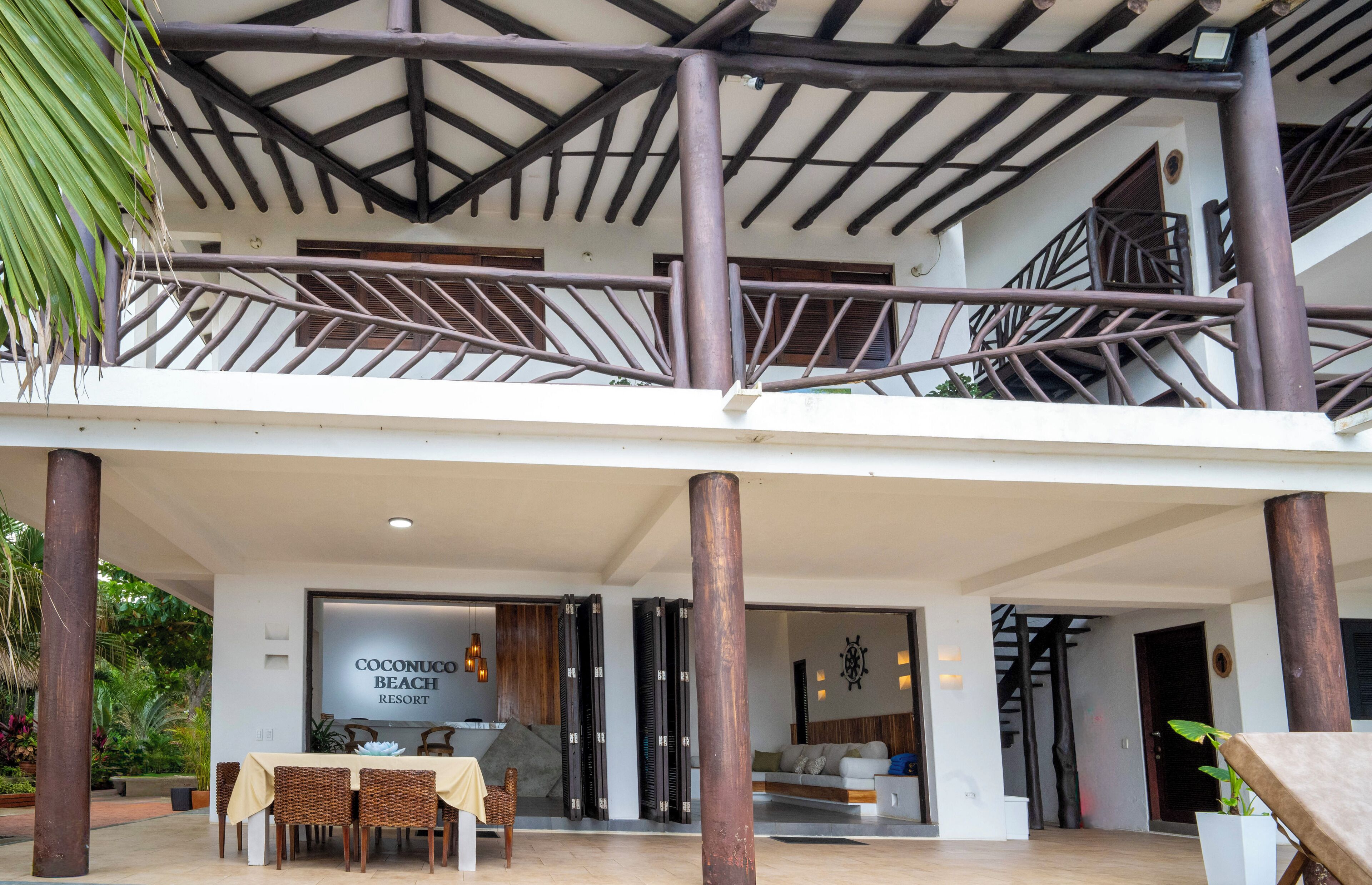 Un edificio de dos plantas con un espacio exterior cubierto, una mesa de comedor con sillas y un letrero que dice "Coconuco Beach Resort".