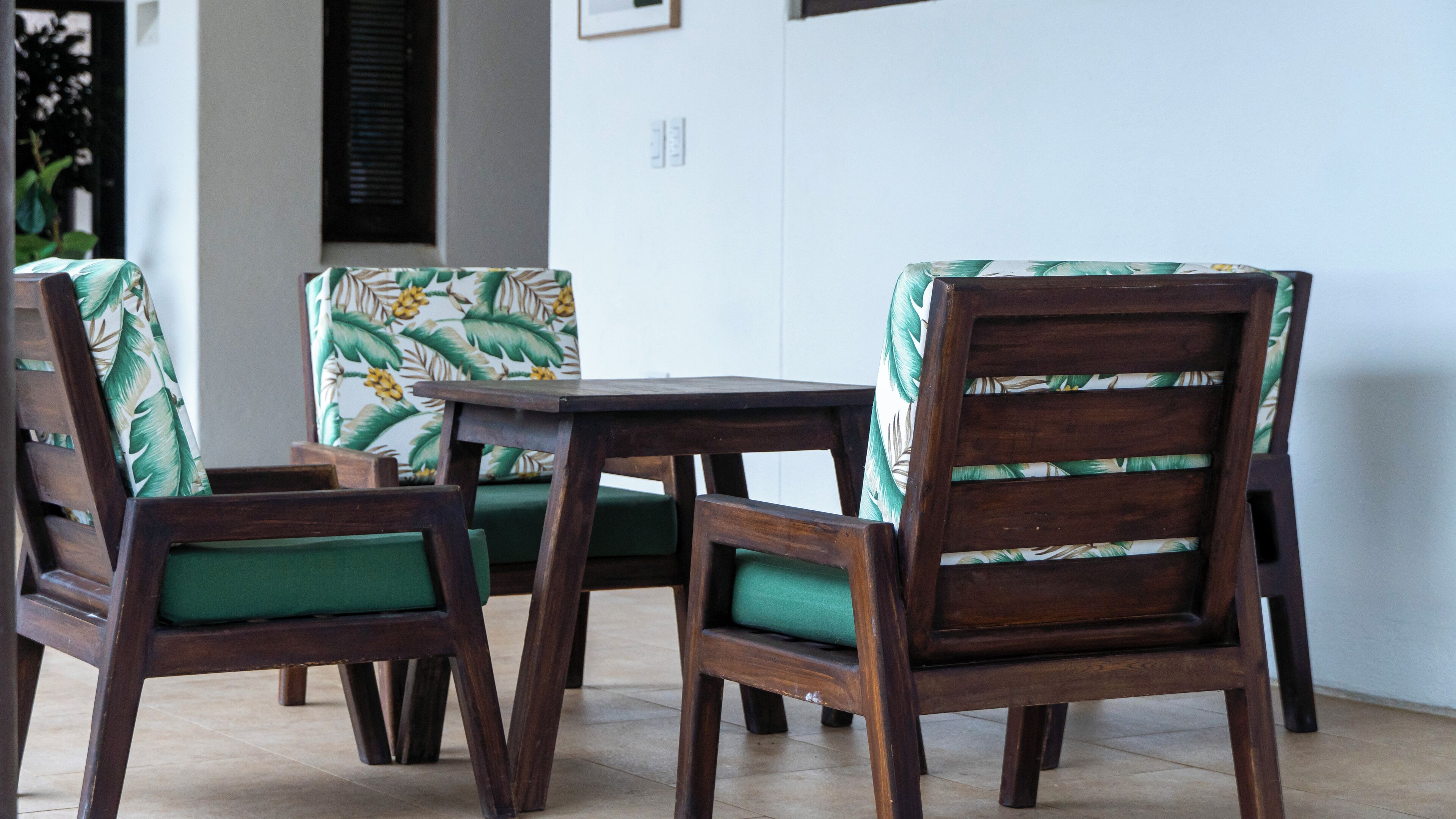 A set of wooden chairs with green cushions and a small table, positioned in a corner with a white wall and a window with blinds.