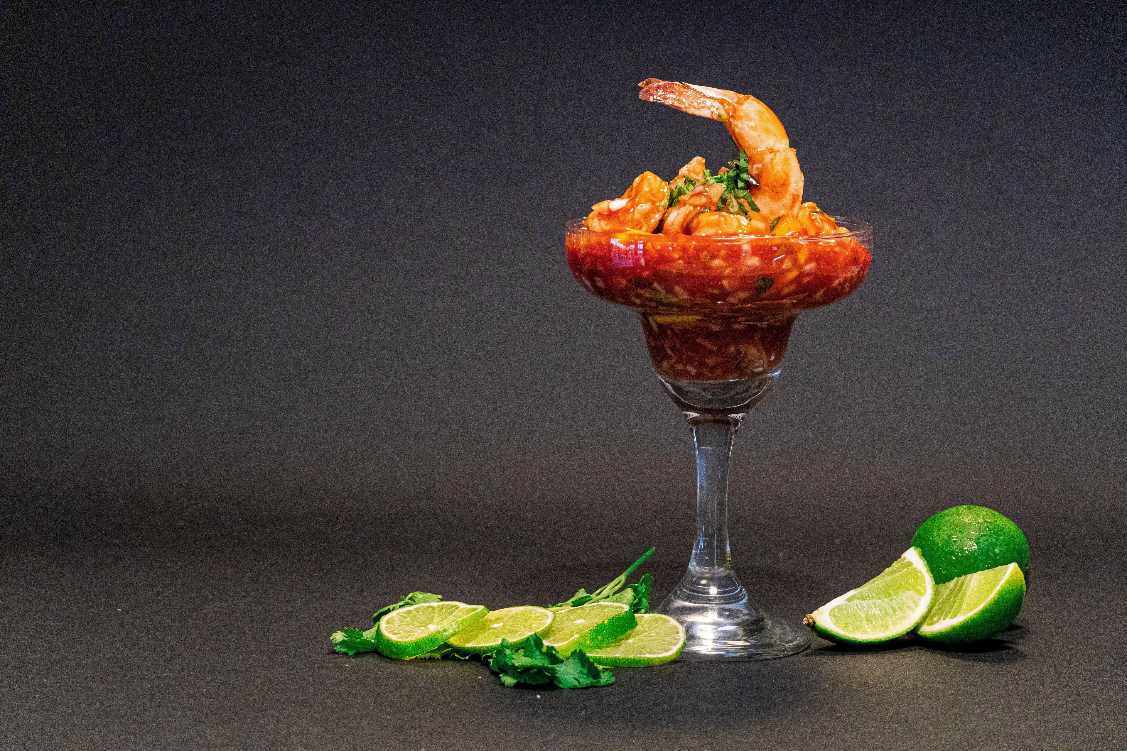 A cocktail in a glass with a shrimp garnish, surrounded by lime slices and herbs.
