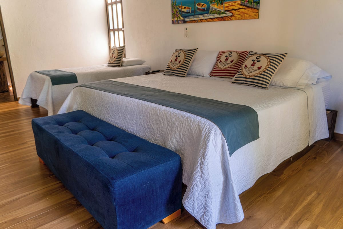 Una cama doble con colcha blanca y almohadas a rayas, un banco tapizado en azul y una mesita de noche con una lámpara.