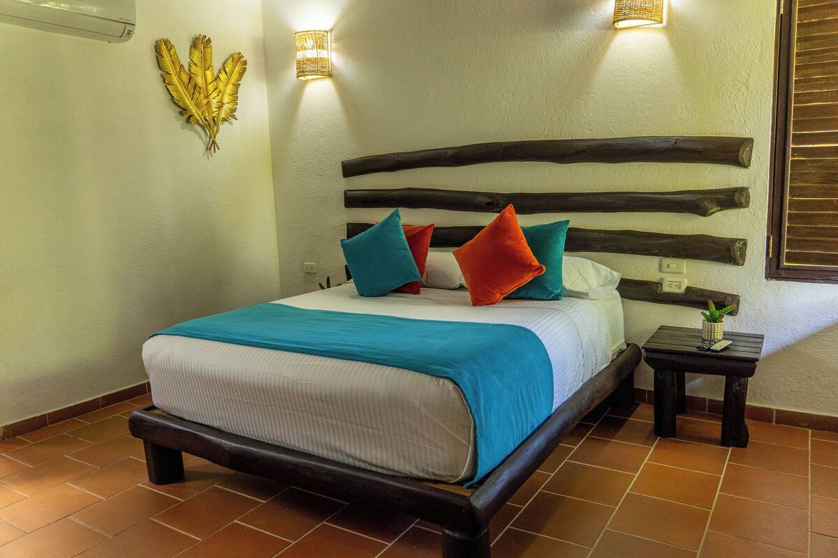 Una cama con cabecera de madera, colcha azul y blanca, y almohadas coloridas. Junto a la cama, una mesita con una planta.