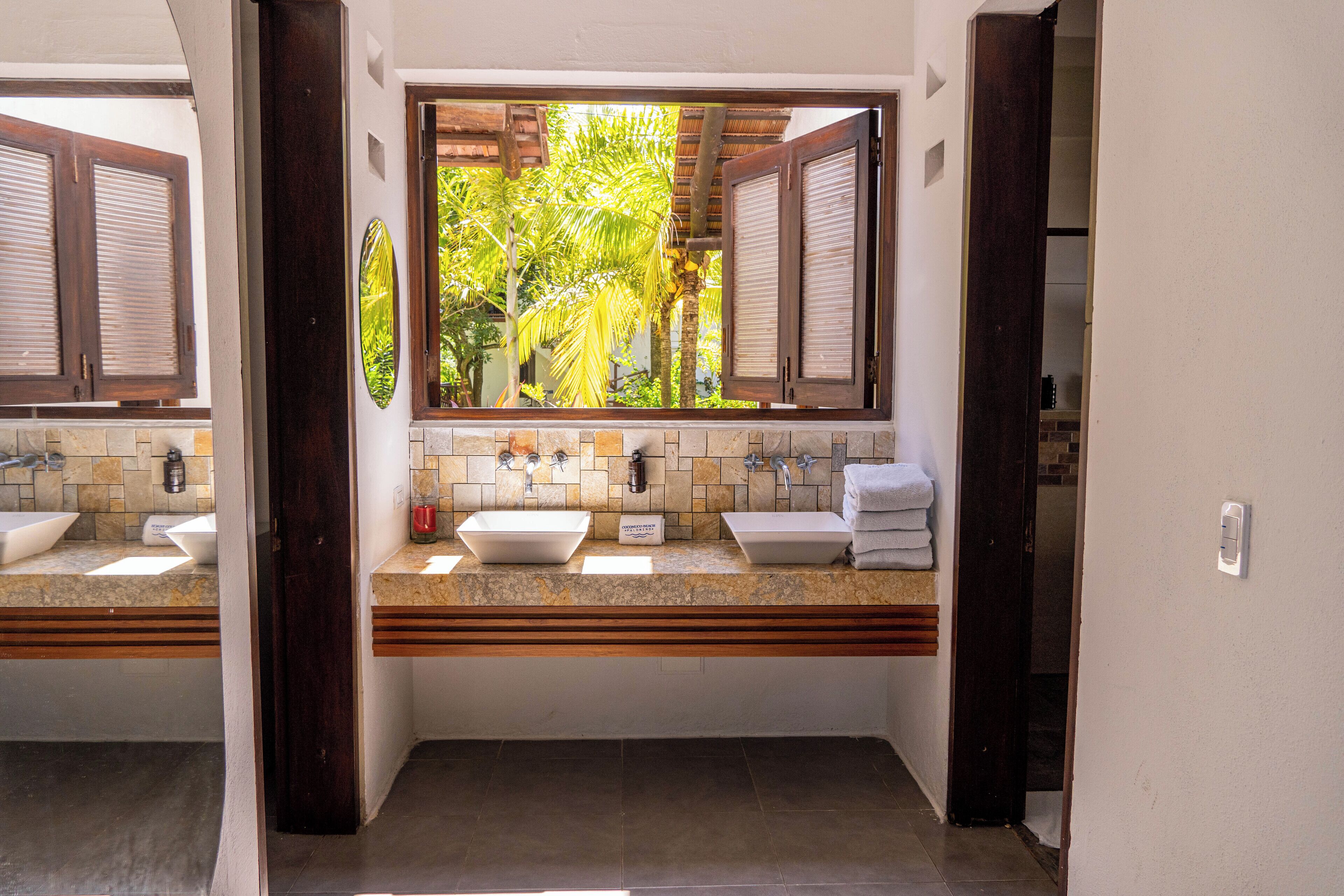 Un baño con dos lavamanos, una ventana con contraventanas y vistas a palmeras.