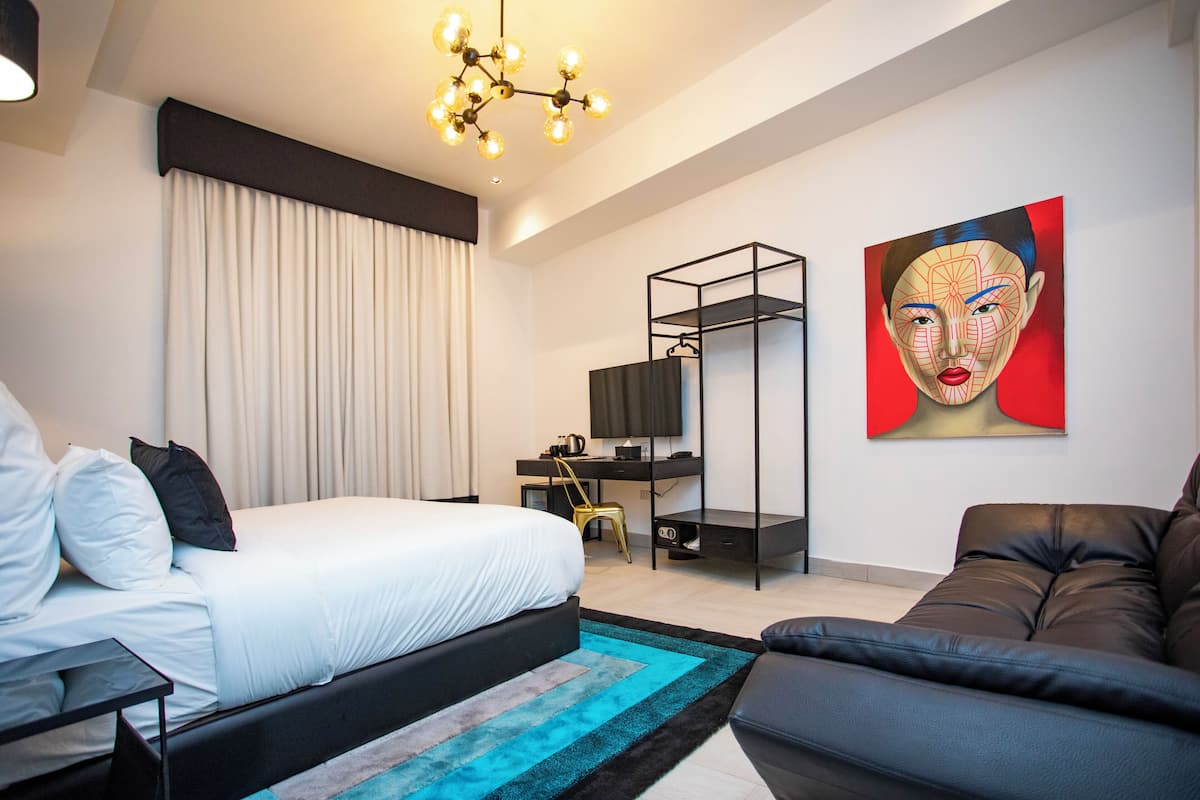 A modern hotel room with a large bed, a desk with a chair, a sofa, and a painting on the wall.