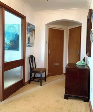 A hallway with a wooden door, a chair, a desk with a telephone, and two paintings on the wall.
