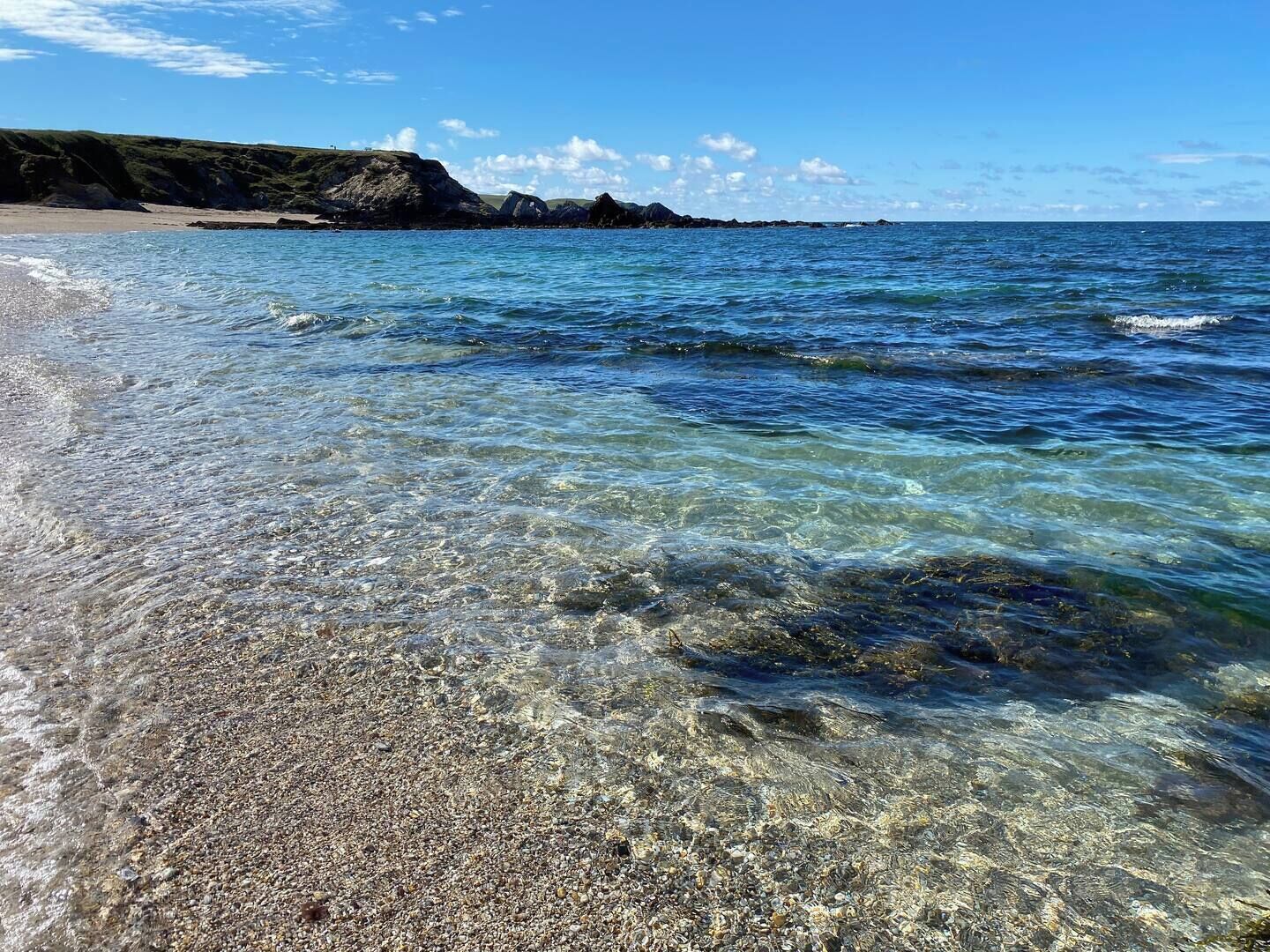 Una platja amb aigües clares i poc profundes, sorra de petits pedres i penya-segats rocosos a la distància.