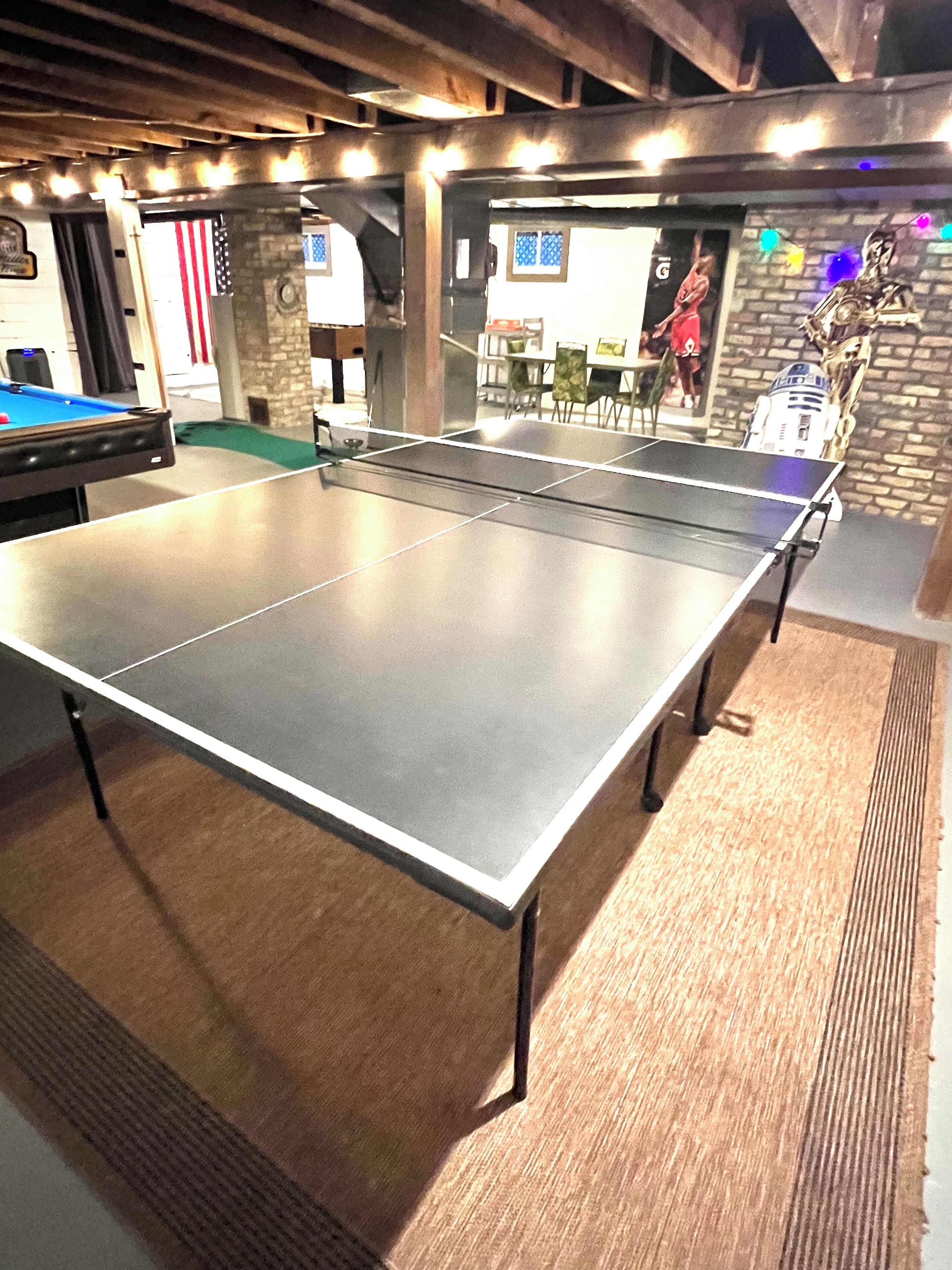 A ping pong table in a room with a pool table, a Star Wars figurine, and a brick wall.