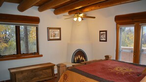 A bedroom with a large bed, a fireplace, wooden beams, and a window with a view of trees. - Centrally Located 3 BR/3 Bath Adobe Home in Taos Mesa NM (El Prado)