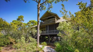 A two-story wooden cabin with a balcony, surrounded by dense greenery and a wooden pathway leading to the entrance. - Canal Front, small dog ok, dock, screened private pool! (St. George Island)