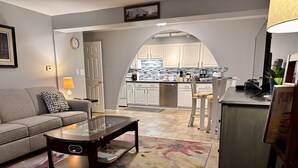Una sala de estar con sofá, mesa de centro y alfombra. Se ve una cocina con armarios y un fregadero al fondo.