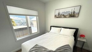 Een slaapkamer met een bed, een raam, een foto aan de muur en een kleine tafel met een rode bloem.