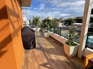 A balcony with a wooden floor, covered grill, potted plants, and a view of the cityscape.