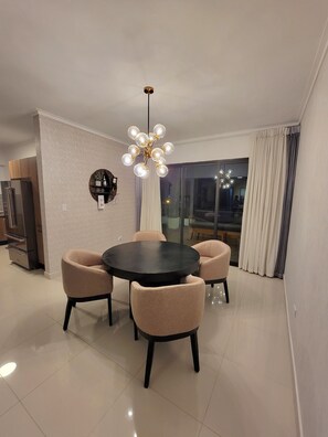A modern dining area with a round table and four chairs, a chandelier, and a view through a glass door.