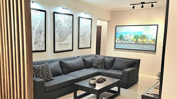 A modern living room with a sectional sofa, a glass coffee table, and framed city maps on the wall. - Brand New luxury condo in Alma Rosa 1, large terrace (Santo Domingo Este)