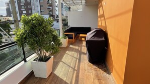 A balcony with wooden flooring, a black sofa, a small table, and a potted plant. - Brand New luxury condo in Alma Rosa 1, large terrace (Santo Domingo Este)