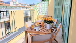 Ein Balkon mit einem Holztisch, gedeckt für ein Frühstück, inklusive Obst, einer Blumenvase und einem Getränk.
