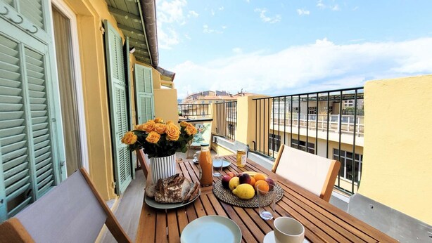 A balcony with a table set for breakfast, including bread, fruit, and a vase of flowers. - Ze Perfect Place - Opera Nice - Apartment 2 Bdrms - AC - Terrace (Nice)