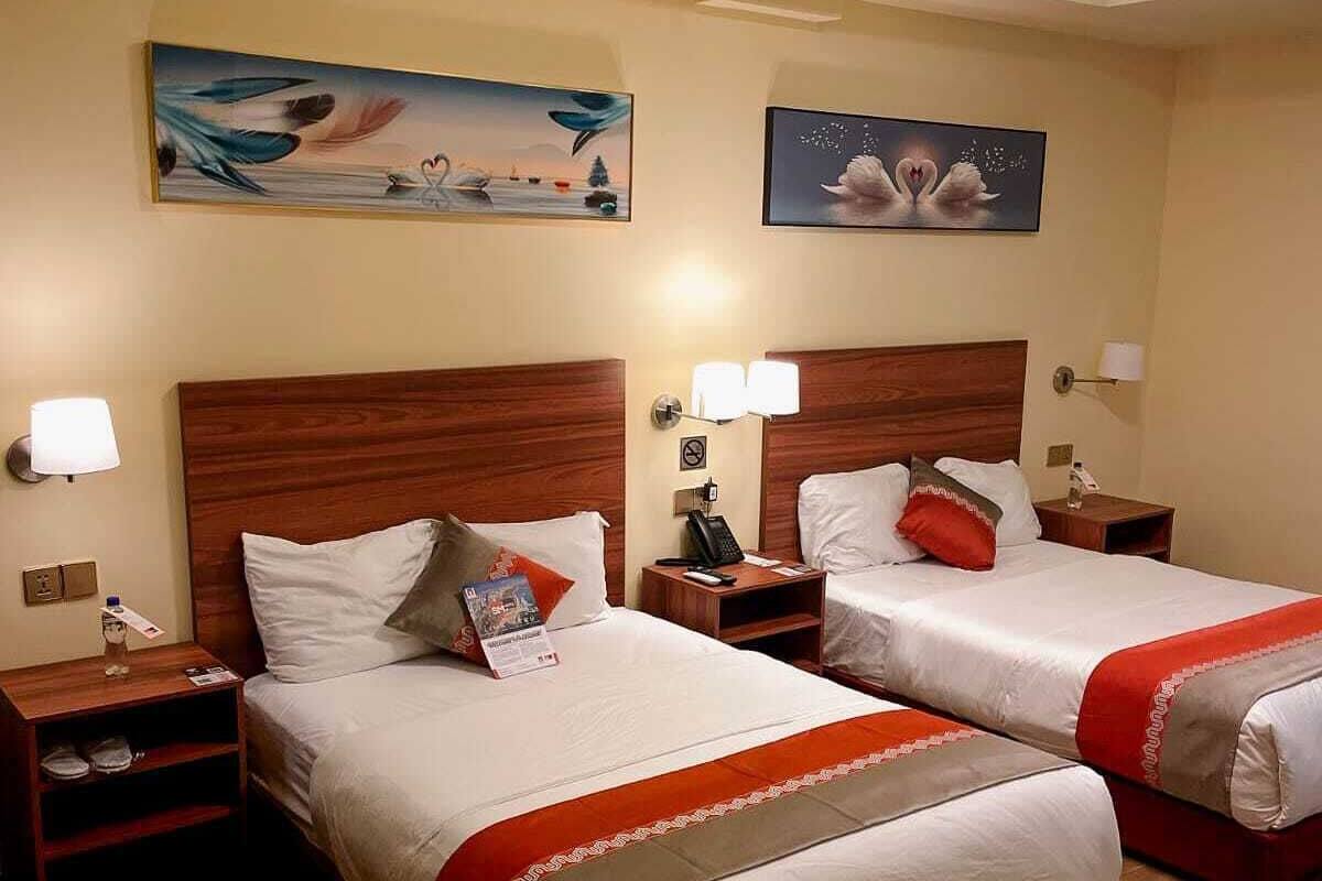 Habitación de hotel con dos camas, cabeceros de madera, mesitas de noche con lámparas y obras de arte en las paredes.
