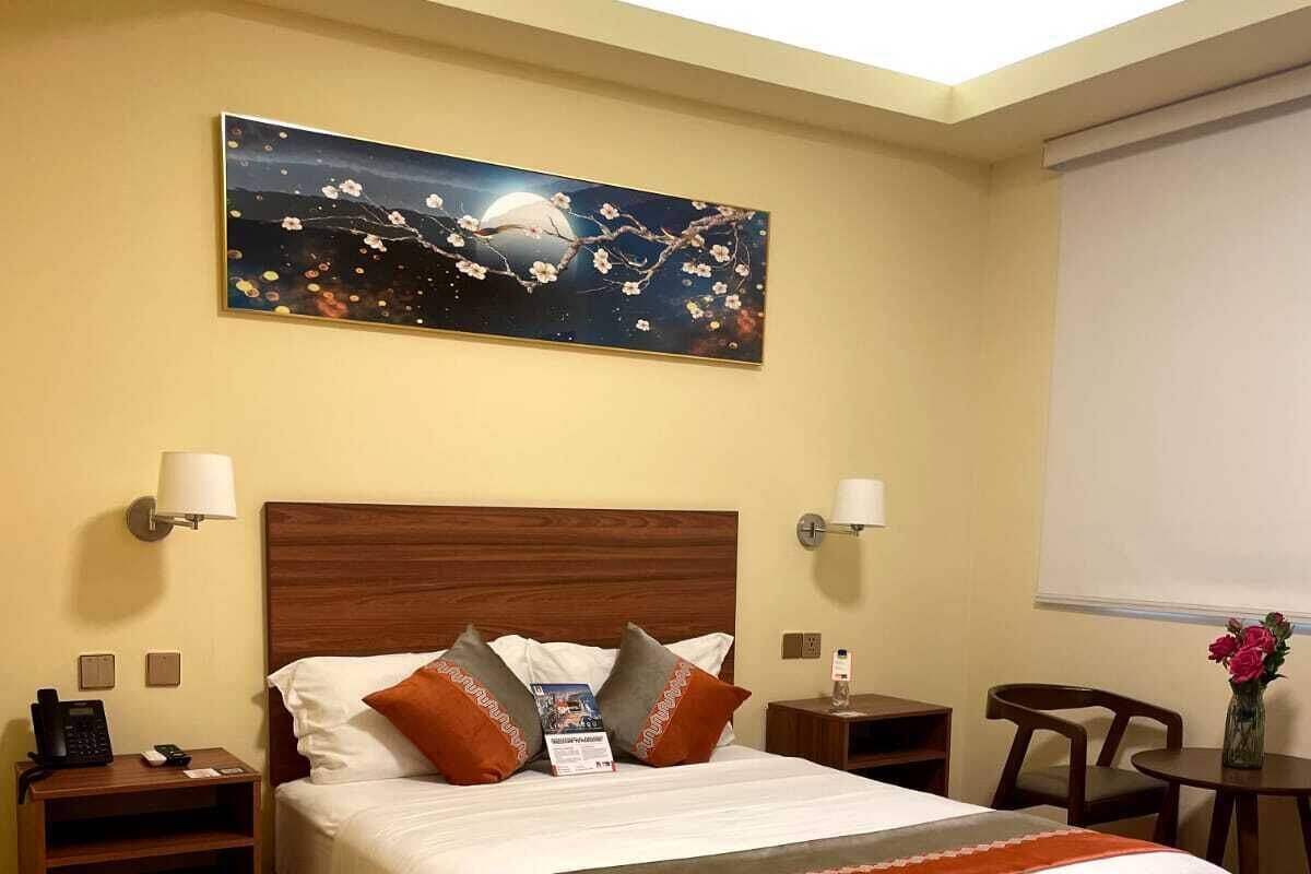 Habitación de hotel con una cama grande, cabecero de madera, dos mesitas de noche con lámparas, una mesita con teléfono, un escritorio con silla y un cuadro en la pared.