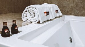 Ett vitt badkar med två flaskor av SM Hotel Burberrys kroppsspray och en hoprullad vit handduk med hotellets logotyp.