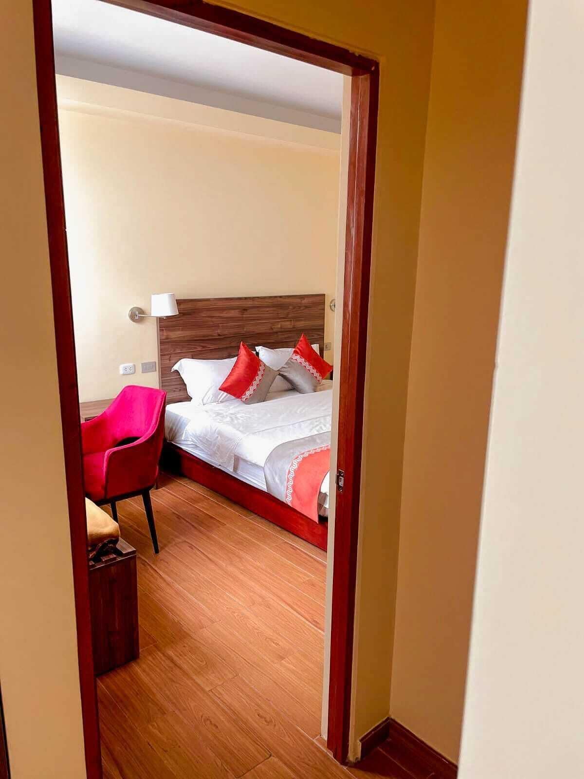 Habitación de hotel con cama de madera, almohadas rojas y blancas, una silla roja y una mesita de noche.