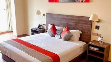Una habitación de hotel con una cama grande, cabecera de madera, dos mesitas de noche, una lámpara de noche, una obra de arte en la pared y una ventana con cortinas.