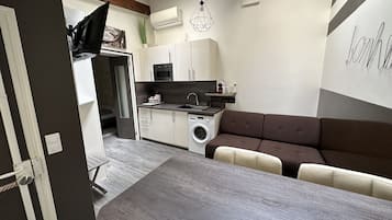 A compact kitchen with a washing machine, a dining area with a table, and a brown sofa.