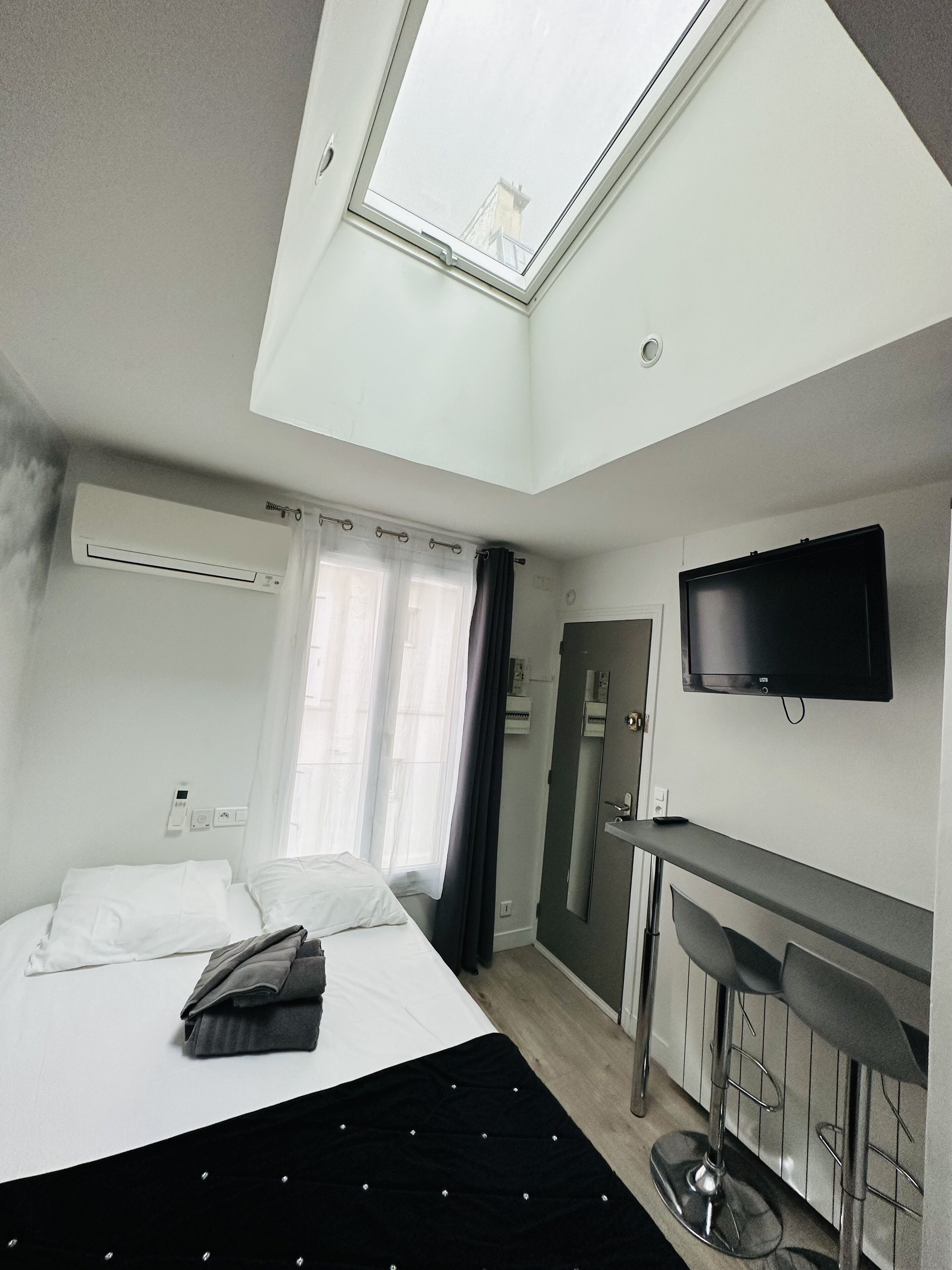 Una habitación de hotel moderna con tragaluz, cama con ropa de cama blanca, manta negra, televisor de pantalla plana y un pequeño bar.