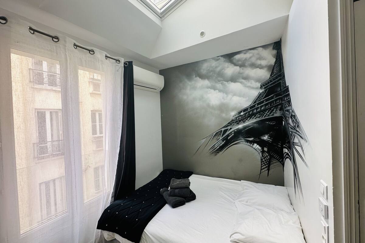 A bedroom with a skylight, a bed with a black quilt, a white wall with a cloud mural, and a window with sheer curtains.