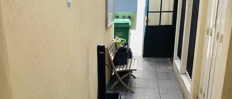 A narrow corridor with tiled flooring, a bench, potted plants, and a green trash bin.