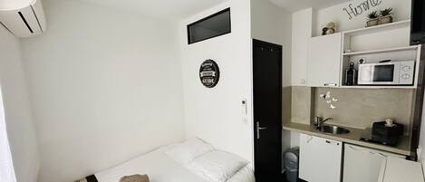 A compact room with a bed, a small kitchenette, a wall-mounted TV, and a door leading to another room.