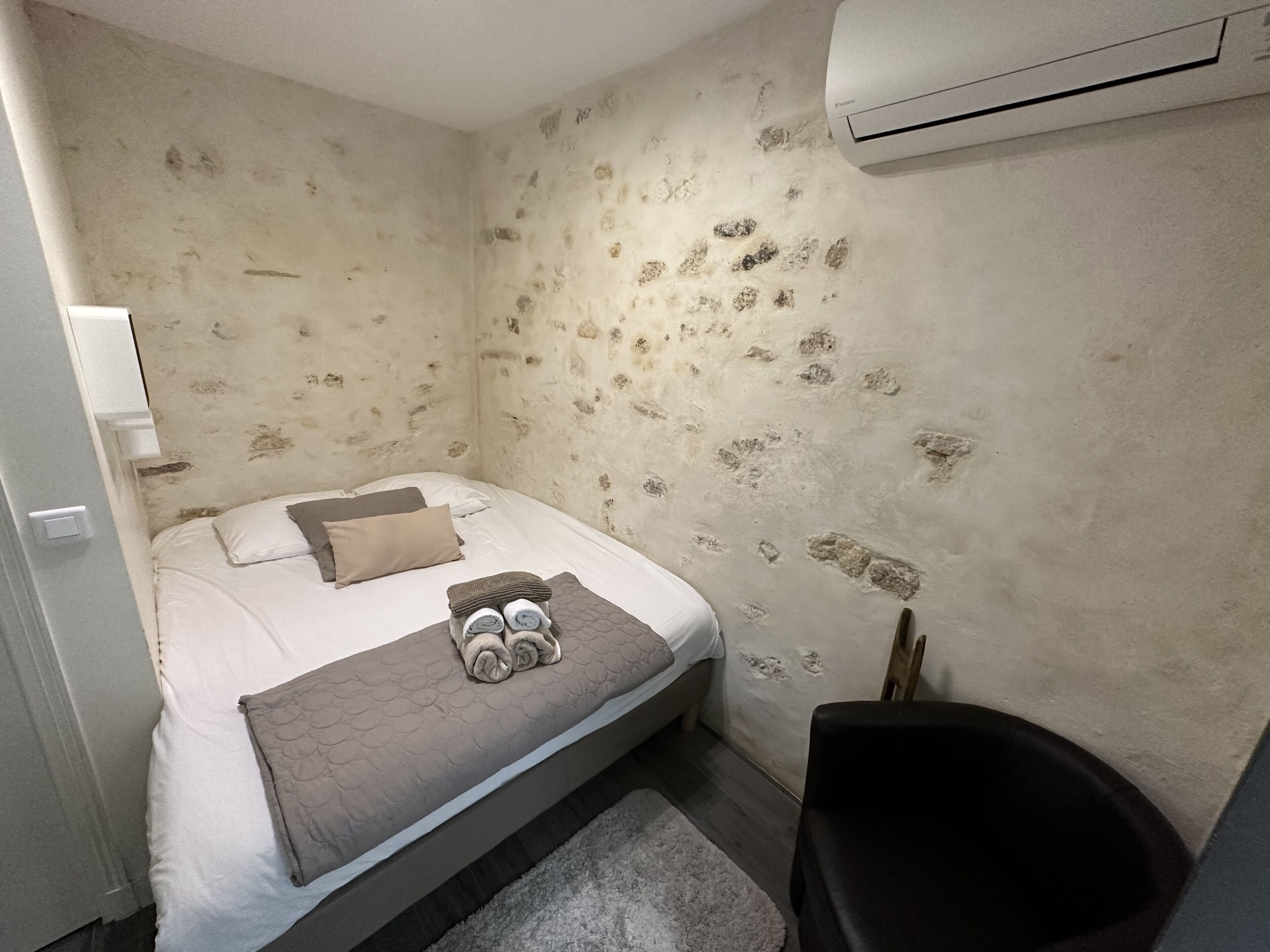 Un dormitorio con cama, almohadas, una colcha y una toalla enrollada sobre la cama. Hay una silla y un equipo de aire acondicionado en la pared.