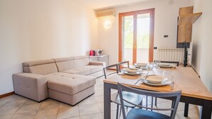 A modern living room with a beige sectional sofa, a dining table set for four, and a kitchen area with a microwave and a red bag on a small table. - Residence San Francesco (Desenzano del Garda)