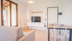 Uma sala de estar moderna e bem iluminada, com um sofá, uma área de jantar com mesa posta para quatro pessoas, uma TV montada acima de um armário branco e uma grande janela com vista para um edifício de tijolos.
