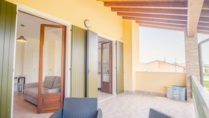 Área externa coberta com sofá e cadeiras, uma porta de madeira e vista para edifícios ao fundo.