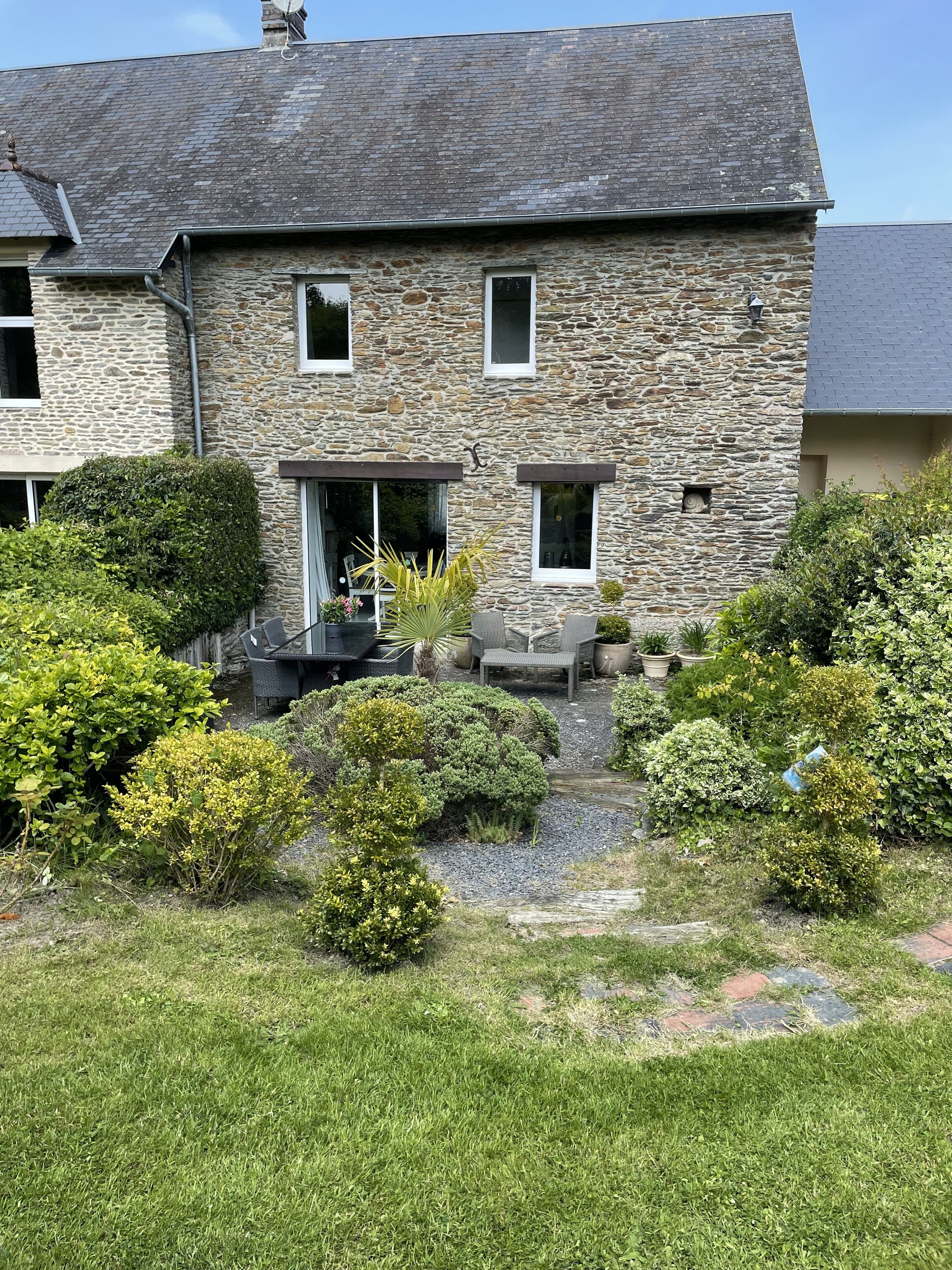 A stone house with a sloped roof, a front porch with outdoor seating, and a well-maintained garden with various shrubs and a small pathway.