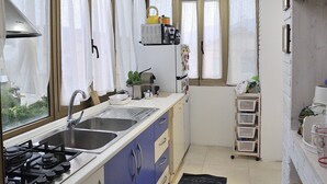 A compact kitchen with a gas stove, sink, and blue cabinets. There are windows with curtains, a small refrigerator, and a shelf with kitchen items. - An apartment nestled in the sun 110 meters from the sea (Scauri)