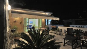 A well-lit outdoor patio area with a dining table and chairs, surrounded by potted plants and a large palm tree. - An apartment nestled in the sun 110 meters from the sea (Scauri)