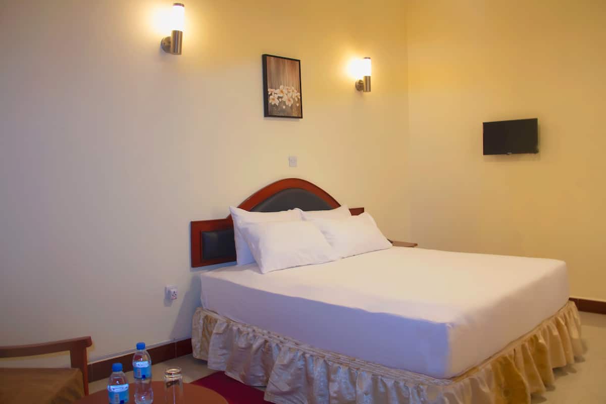 A neatly made bed with white linens, a wooden headboard, and a decorative ruffled skirt. There are two wall-mounted lights and a flat-screen TV on the wall.