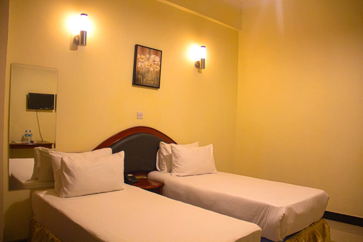 A hotel room with two single beds, a wooden headboard, a bedside table with a lamp, a flat-screen TV, and a painting on the wall.