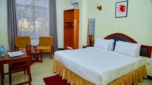 Habitació d'hotel amb llit, escriptori i cadira, armari i bany visible a través d'una finestra amb cortines.