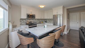 A modern kitchen with white cabinets, a stainless steel refrigerator, a microwave, and a built-in oven. There is a marble island with bar stools and a window with blinds. - Newly renovated with Lakeviews, big yard, minutes to Silver Star/Golf/Wineries. (Vernon)