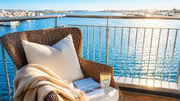 A wicker chair with a cushion and a blanket, a glass of water with a lemon slice, and a menu on a balcony overlooking a marina. - Dog-friendly Baltic Sea vacation apartment with a wonderful view of the harbor basin (Olpenitz)