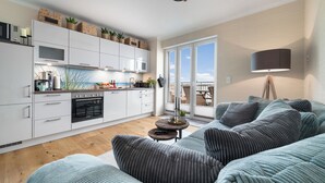 A modern kitchen with white cabinets, a built-in oven, and a microwave. There is a dining area with a table and chairs, and a large window offering a view of the outdoors. - Dog-friendly Baltic Sea vacation apartment with a wonderful view of the harbor basin (Olpenitz)