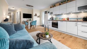 A modern kitchen with white cabinets, a dining area with a blue sofa, and a flat-screen TV mounted on the wall. - Dog-friendly Baltic Sea vacation apartment with a wonderful view of the harbor basin (Olpenitz)