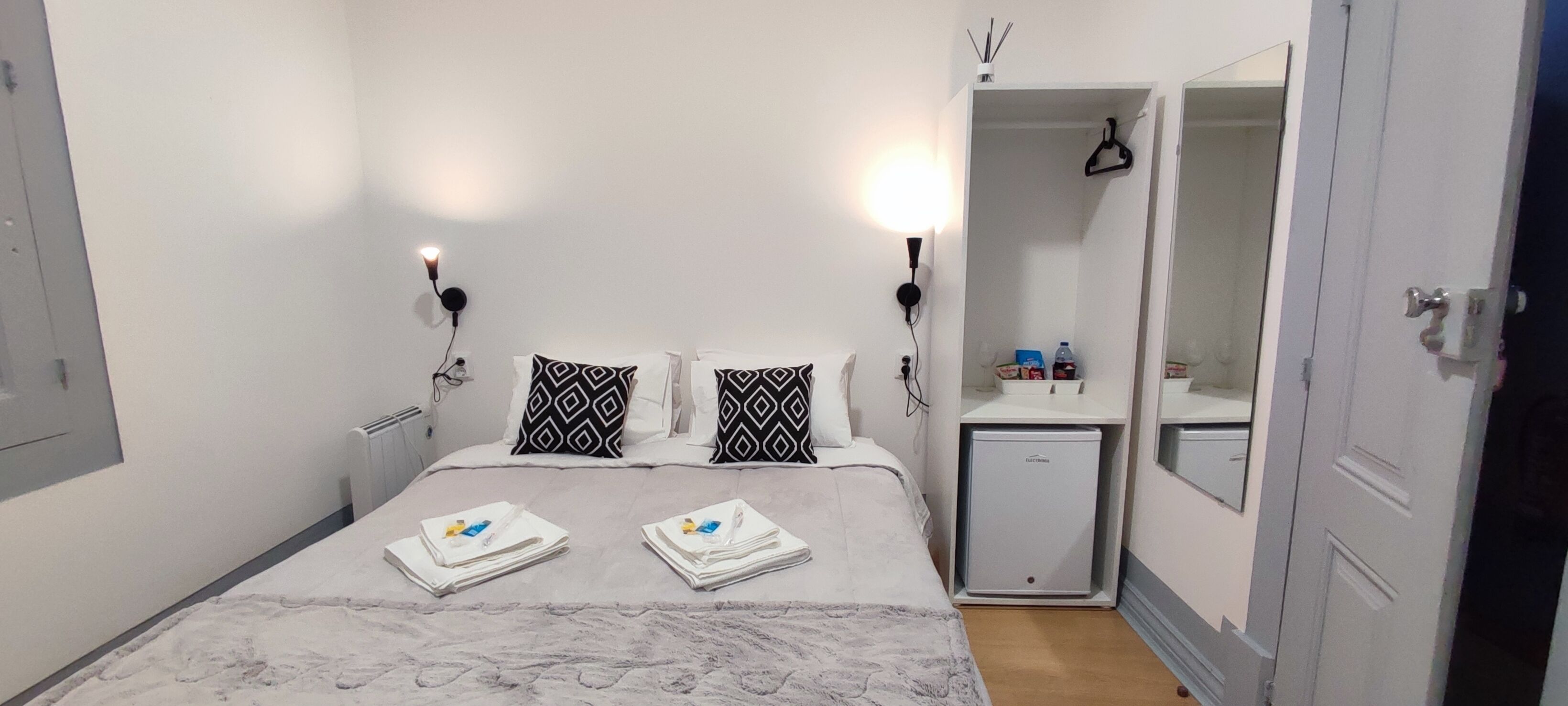 Una habitación de hotel con cama, dos lámparas de noche, un pequeño armario blanco, un espejo y una puerta.