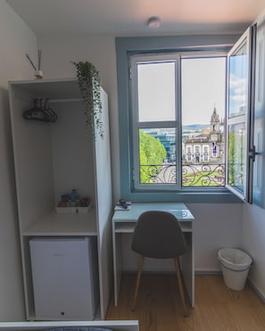 Een kleine kamer met een bureau, een stoel, een radiator, een prullenbak en een raam met uitzicht op een gebouw en bomen.